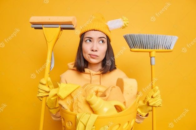 Tired thoughtful asian woman does laundry at home holds cleaning tools busy with cleanup going to sweep floor wash toilet wears hat sweatshirt and rubber gloves isolated over yellow background | Premium Photo