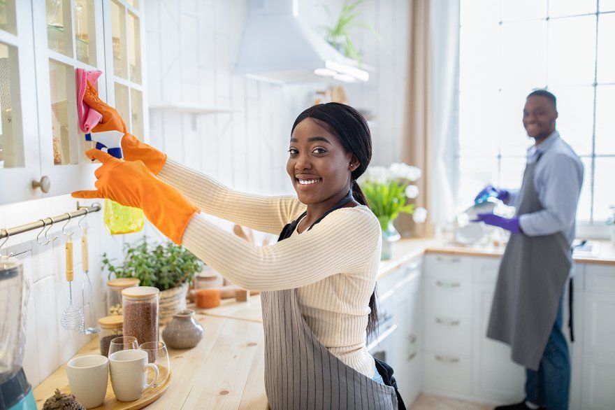 Happy black lady cleaning kitchen cabinet, her husband washing dishes on backgr